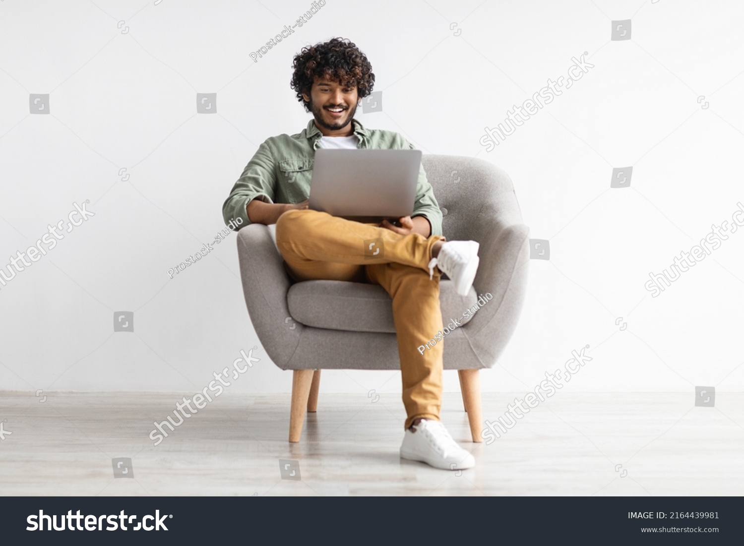 Joyful young indian guy in casual sitting in armchair, chilling with modern laptop, gambling or gaming online, surfing on Internet, white studio background, full length shot, copy space #2164439981