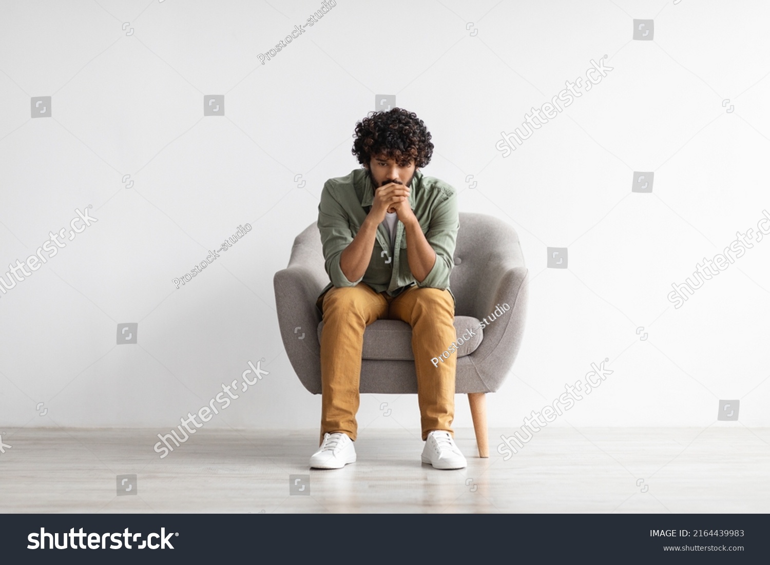 Depressed curly bearded young indian guy in casual outfit sitting in armchair over white studio background, looking down, thinking about something, suffering from loneliness, copy space, full length #2164439983