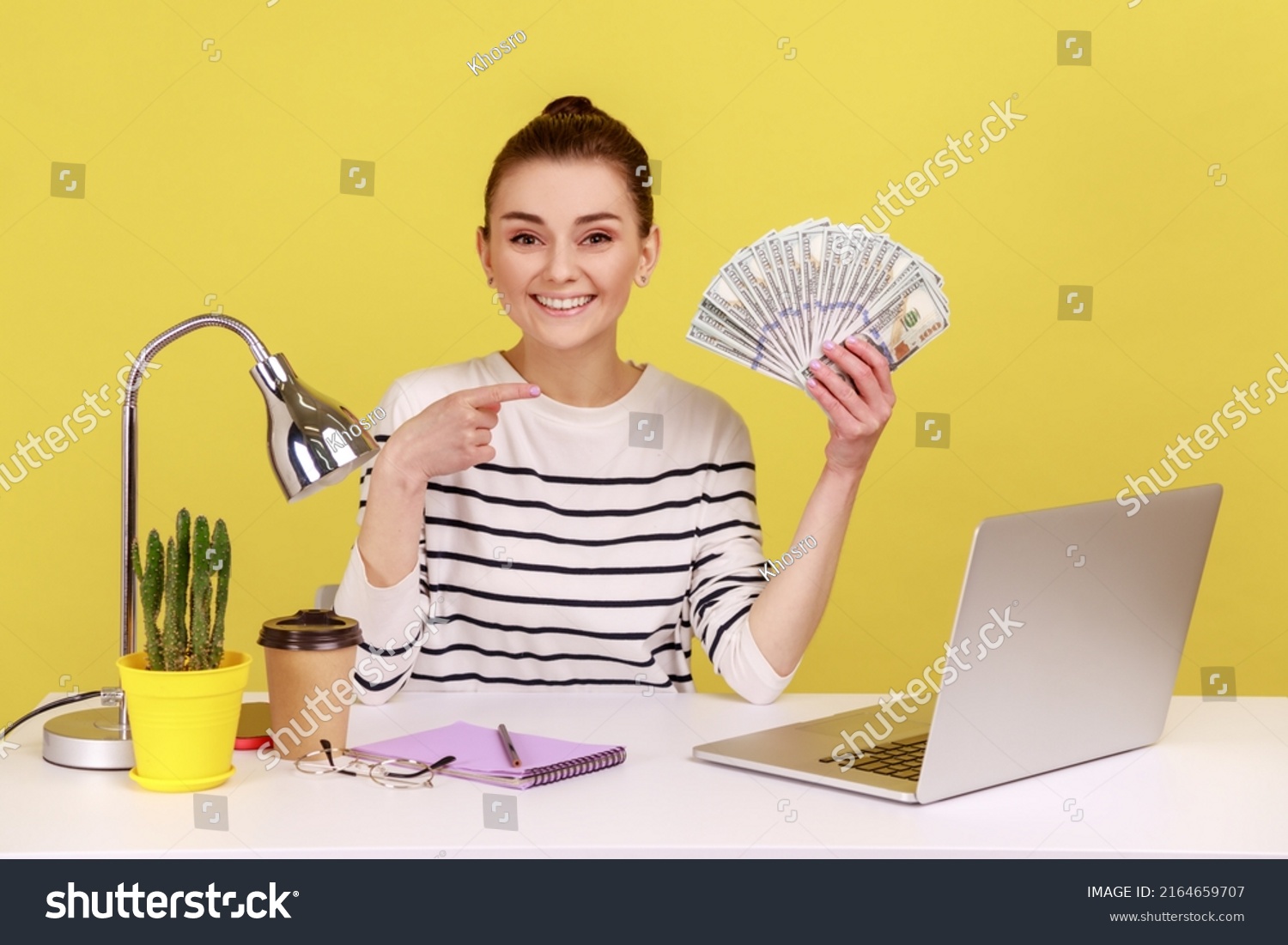 Positive young woman pointing finger at hundred dollar bills, holding sitting at workplace with laptop, high salary, bonuses and perks. Indoor studio studio shot isolated on yellow background. #2164659707