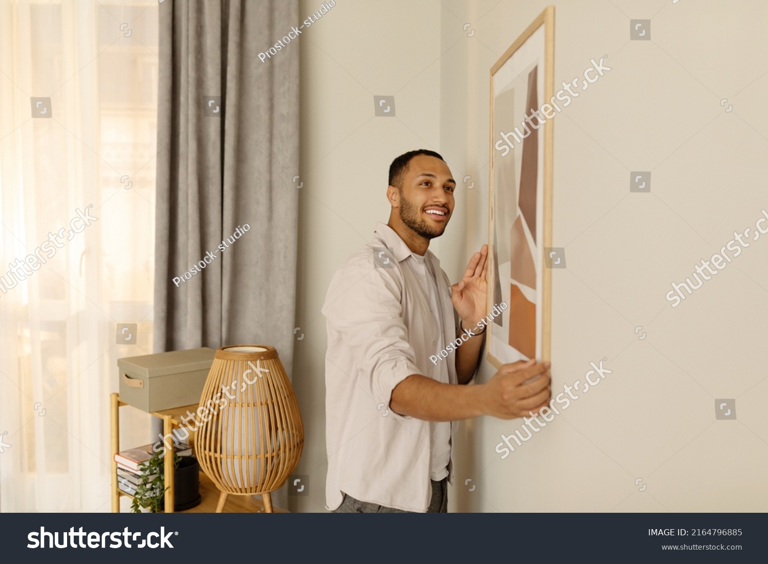 Smiling young black man putting picture frame, hanging painting on wall, empty space. African American male interior designer decorating new modern stylish apartment. Home interior and domestic decor #2164796885