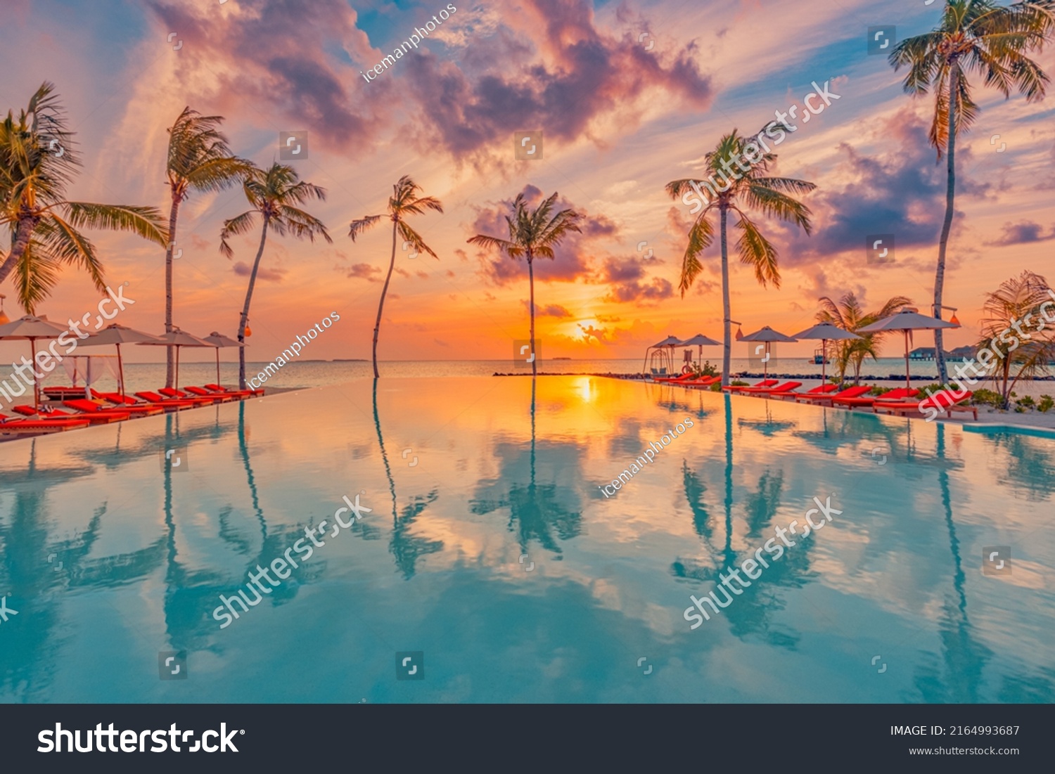 Tropical sunset over outdoor infinity pool in summer seaside resort, beach landscape. Luxury tranquil beach holiday, poolside reflection, relaxing chaise lounge romantic colorful sky, chairs umbrella #2164993687