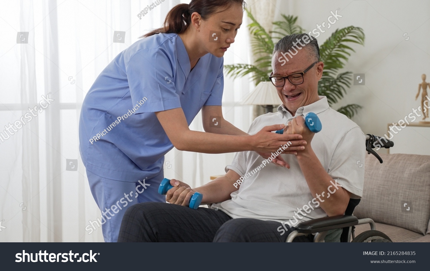 asian paralyzed elderly man sitting in wheelchair and going through physical therapy with personal care attendant’s support. he does weight lifting using dumbbells at home #2165284835