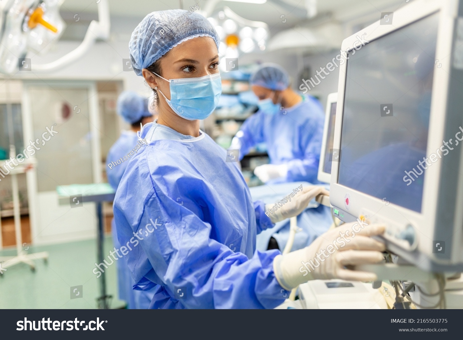 Anesthetist Working In Operating Theatre Wearing Protecive Gear checking monitors while sedating patient before surgical procedure in hospital #2165503775