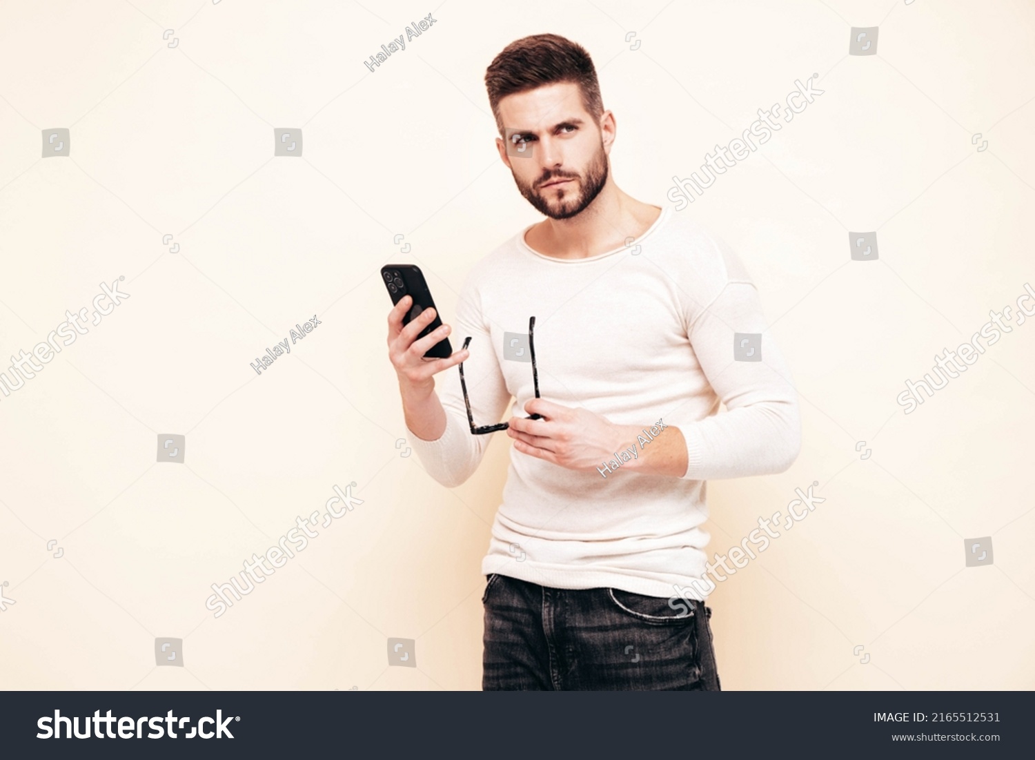 Handsome confident model.Sexy stylish man dressed in sweater and jeans. Fashion hipster male posing near white wall in studio. Holding smartphone. Looking at cellphone screen. Using apps #2165512531