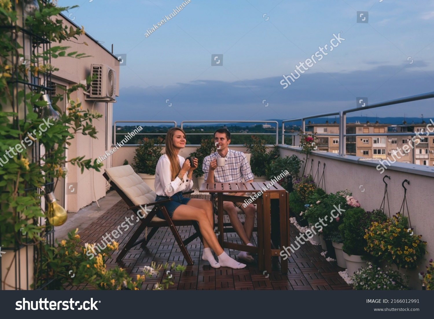 Young couple relaxing outdoors on urban rooftop garden with blooming flowers. Man and woman in casual clothes enjoying sunset and drinking tea or coffee at apartment balcony terrace with city view. #2166012991