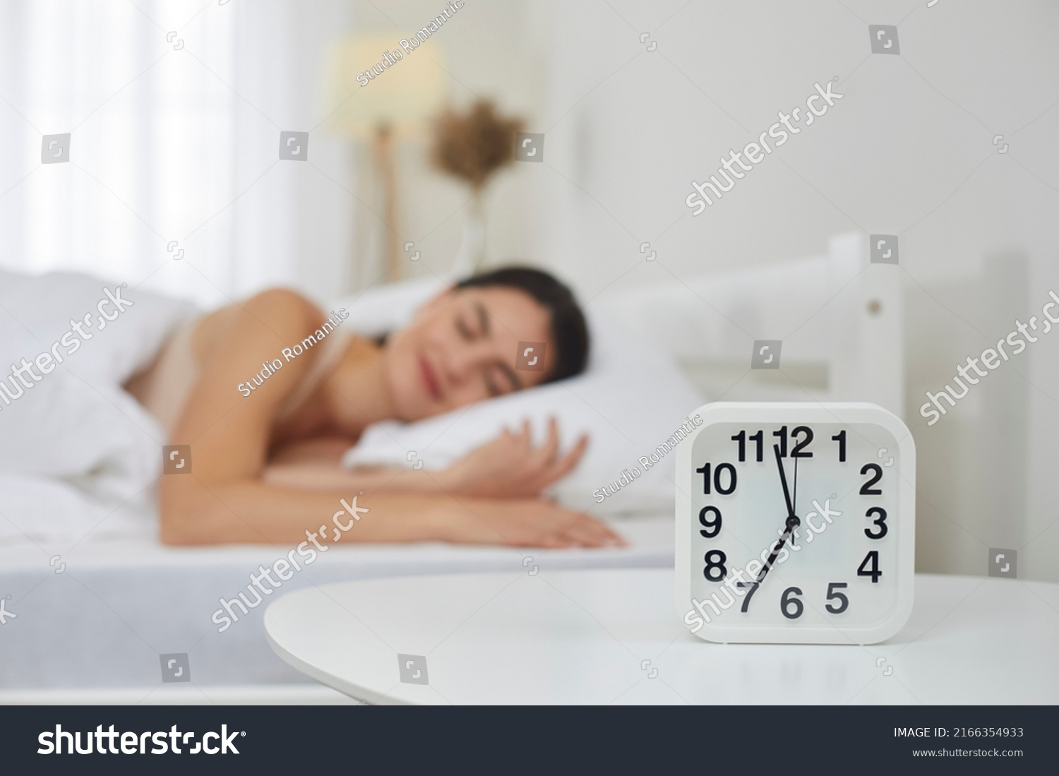 Close up of white plastic alarm clock set for 7 AM on bedside table in bedroom, with happy peaceful tranquil young woman sleeping on bed in blurred background. Sleep, morning, daily routine concepts #2166354933