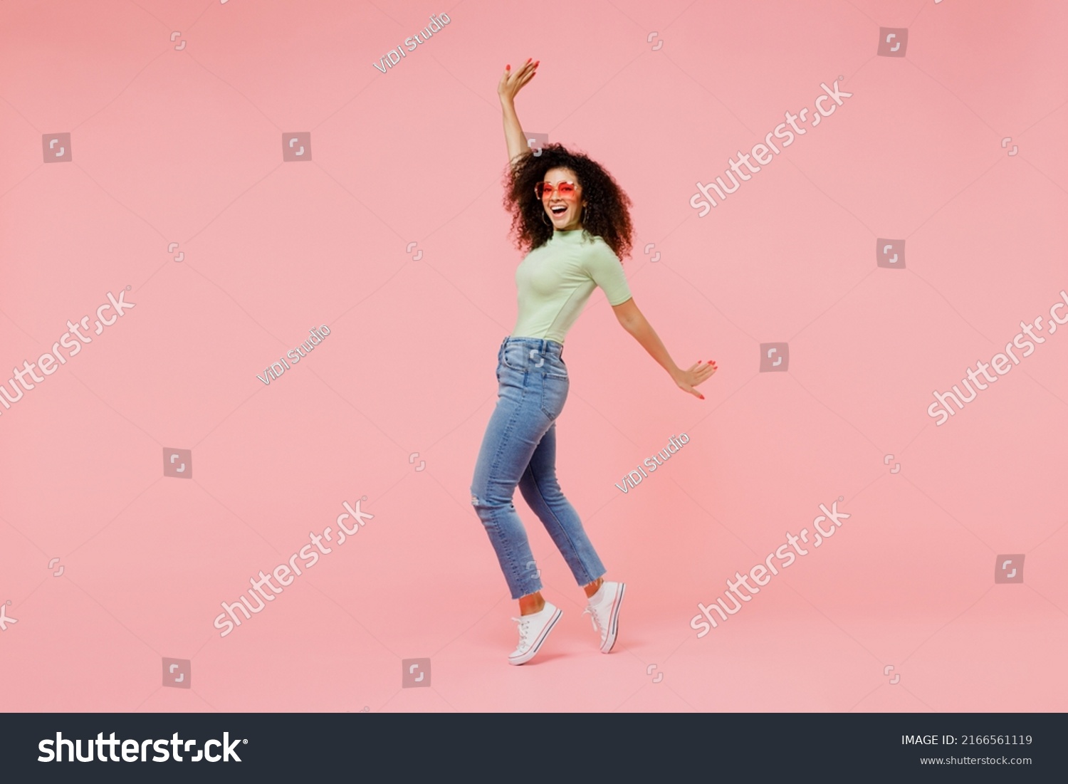 Full size body length young curly latin woman 20s wears casual clothes sunglasses stand on toes dance lean back have fun spreading hands isolated on plain pastel light pink background studio portrait #2166561119