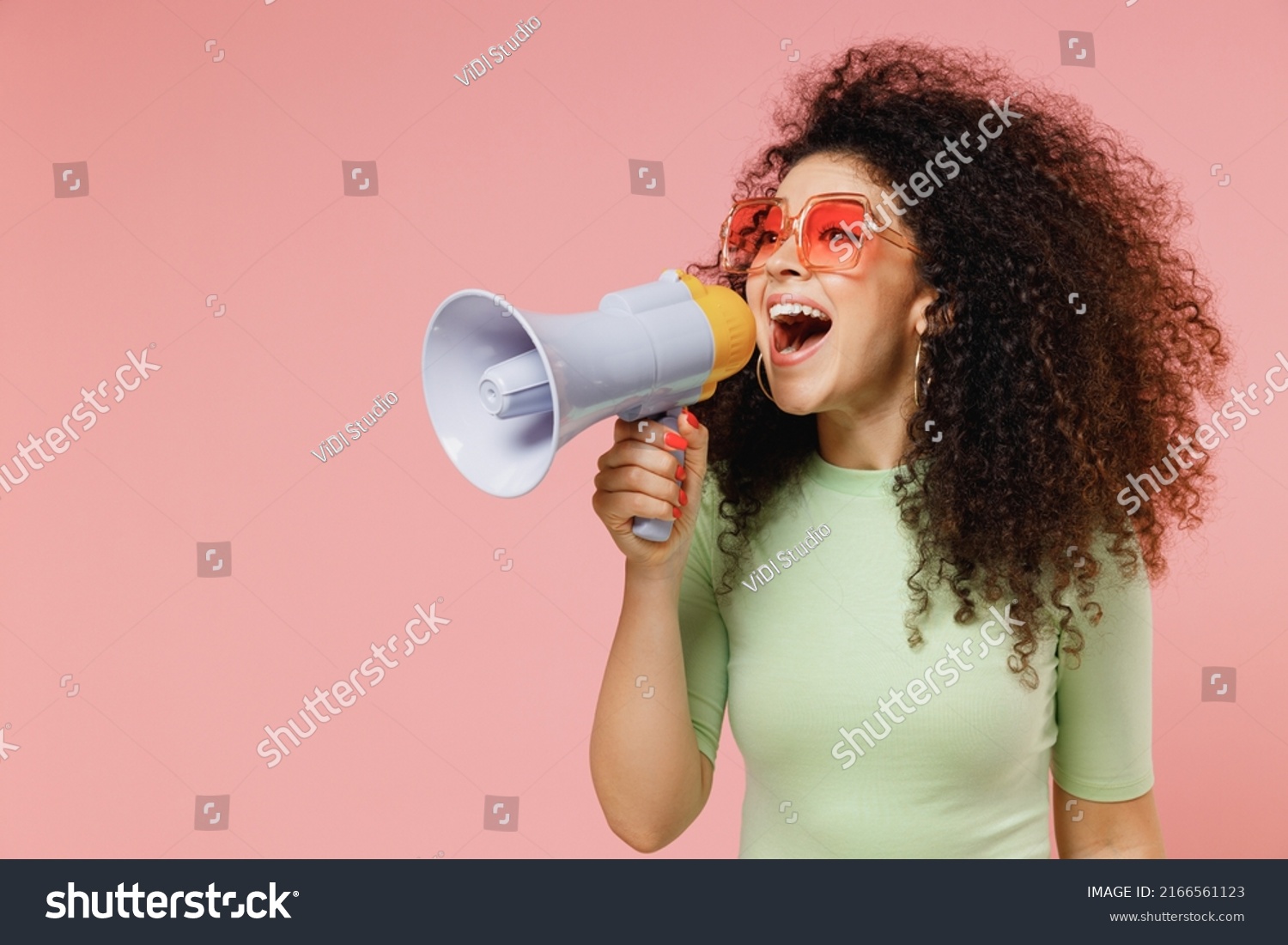 Exultant happy vivid young curly latin woman 20s wears mint t-shirt sunglasses hold scream in megaphone announces discounts sale Hurry up isolated on plain pastel light pink background studio portrait #2166561123