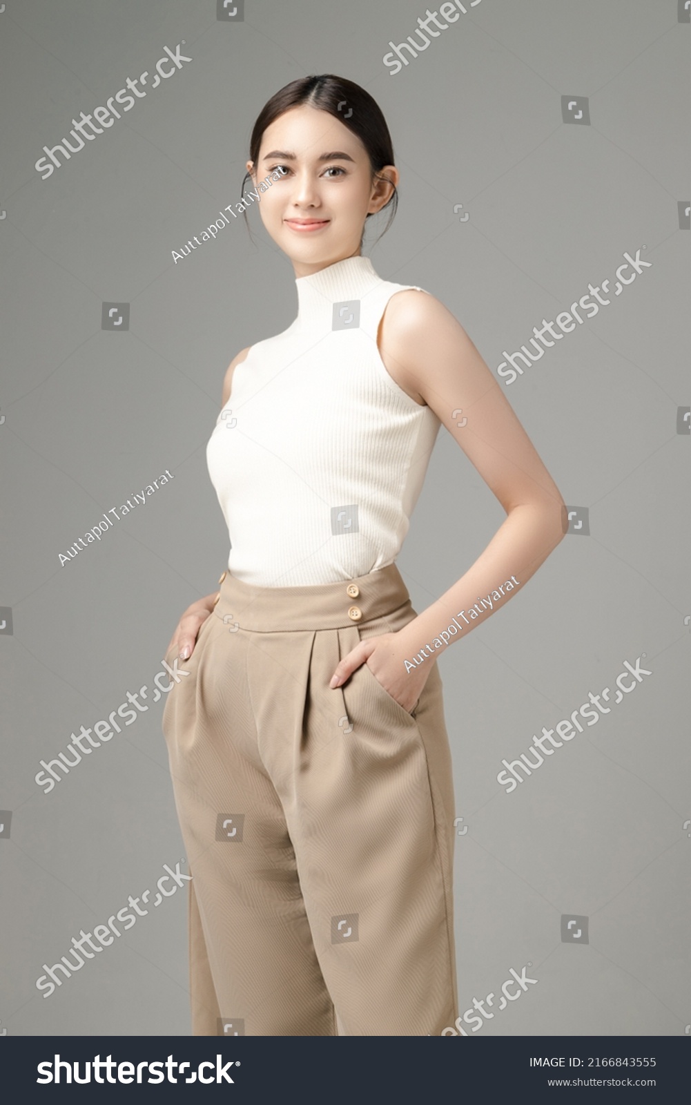 Confident Asian woman puts her hands in pocket and looks at the camera isolated over grey background. Portrait of pretty girl in studio. Perfect body. #2166843555
