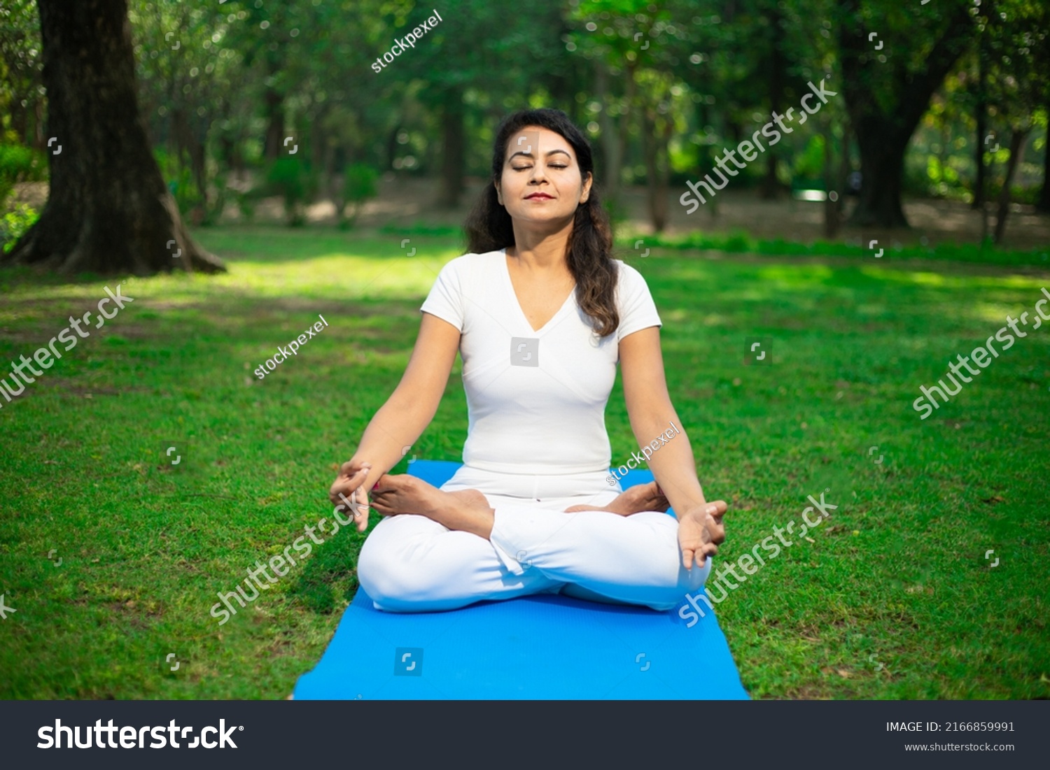 Beautiful young indian woman wearing white cloths doing yoga meditation outdoor in nature Brunette female breathing exercise in park alone, fitness,  Mental health, Peaceful mind,. Copy space.  #2166859991
