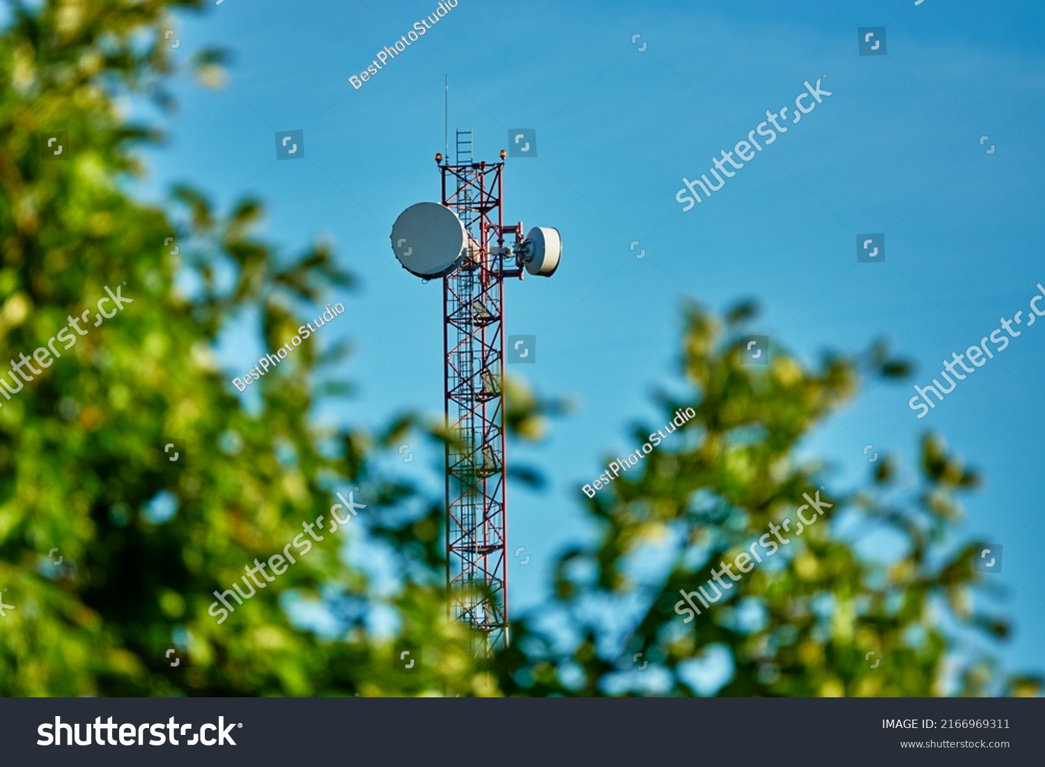 Mast with cell phone antennas against blue sky. Tree with green leaves. #2166969311