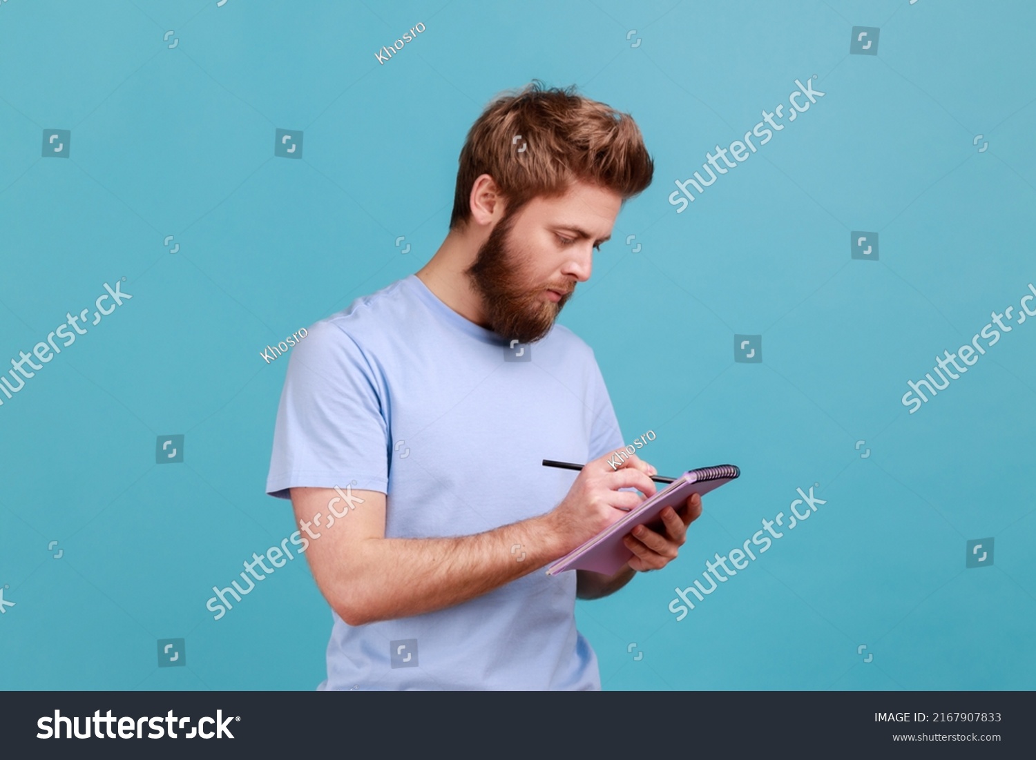 Portrait of bearded man making notes in paper notebook, writing business idea, future plans, checking appointment in schedule diary. Indoor studio shot isolated on blue background. #2167907833