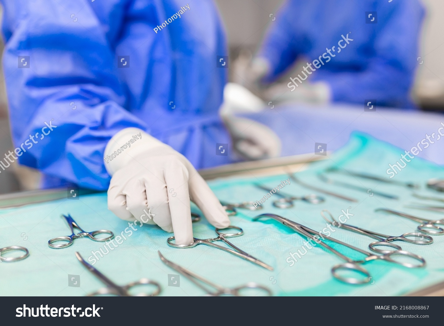 Nurse hand taking surgical instrument for group of surgeons at background operating patient in surgical theatre. Steel medical instruments ready to be used. Surgery and emergency concept #2168008867