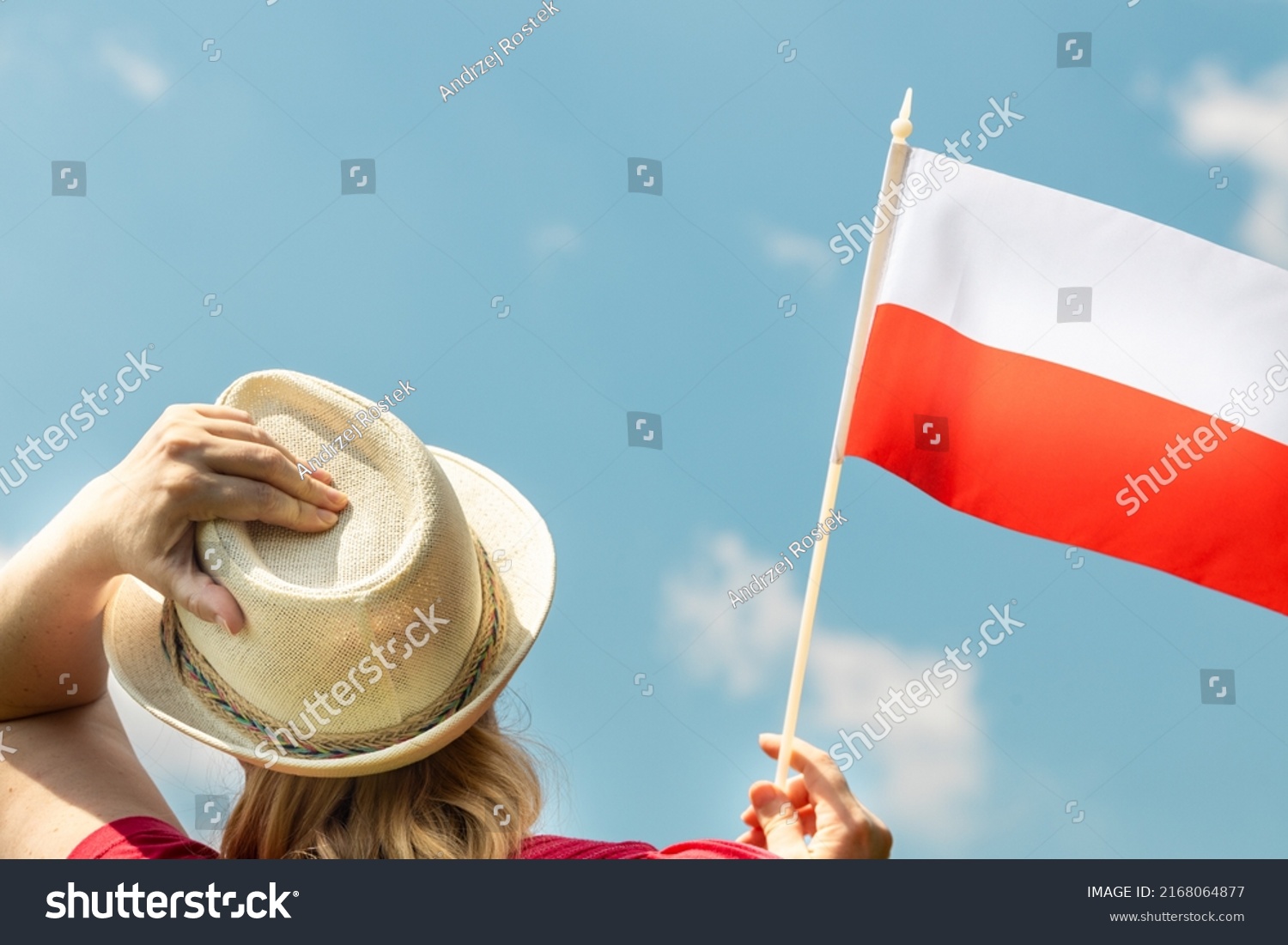 A young woman proudly holds the Polish flag above her head, against the sky with her other hand holding her hat, Concept of national holidays and patriotic events #2168064877