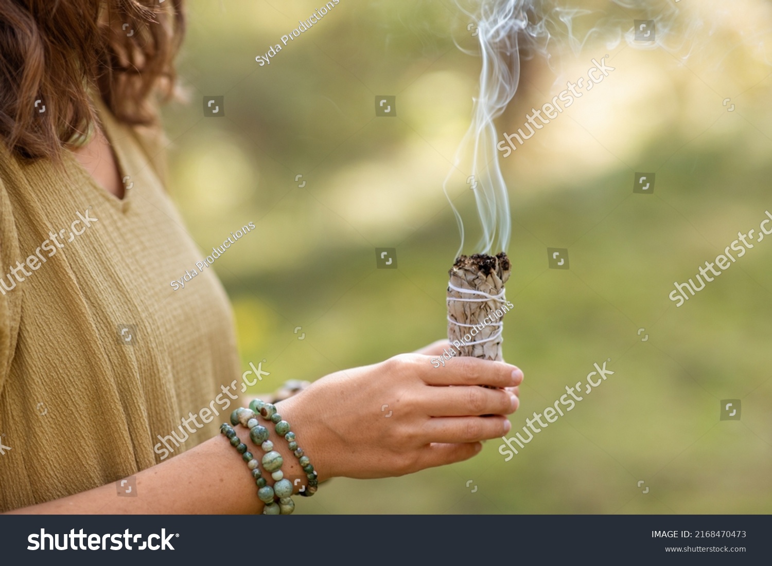 occult science and supernatural concept - close up of woman or witch with smoking white sage performing magic ritual in forest #2168470473