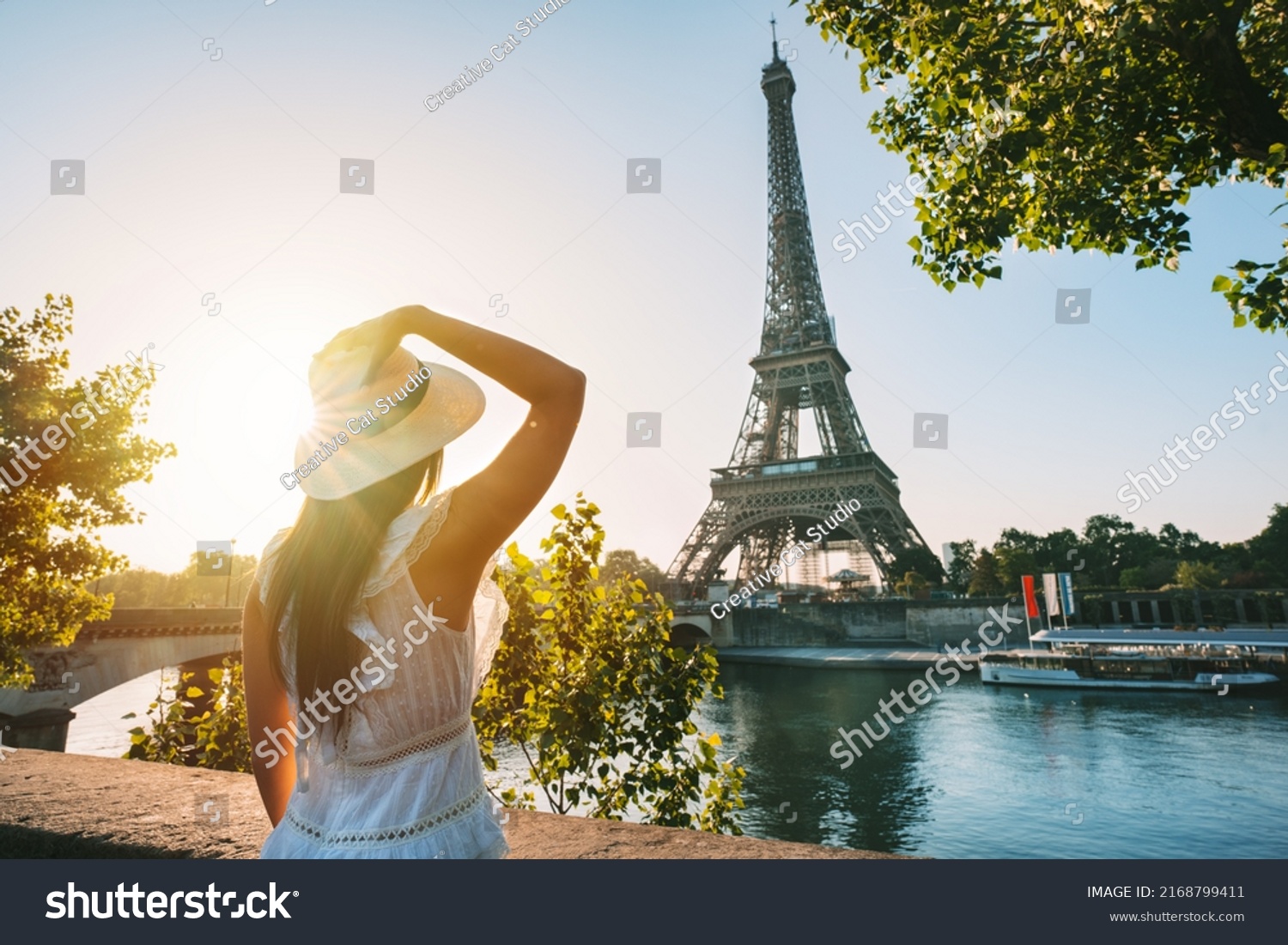 Young woman tourist in sun hat and white dress standing in front of Eiffel Tower in Paris at sunset. Travel in France, tourism concept #2168799411