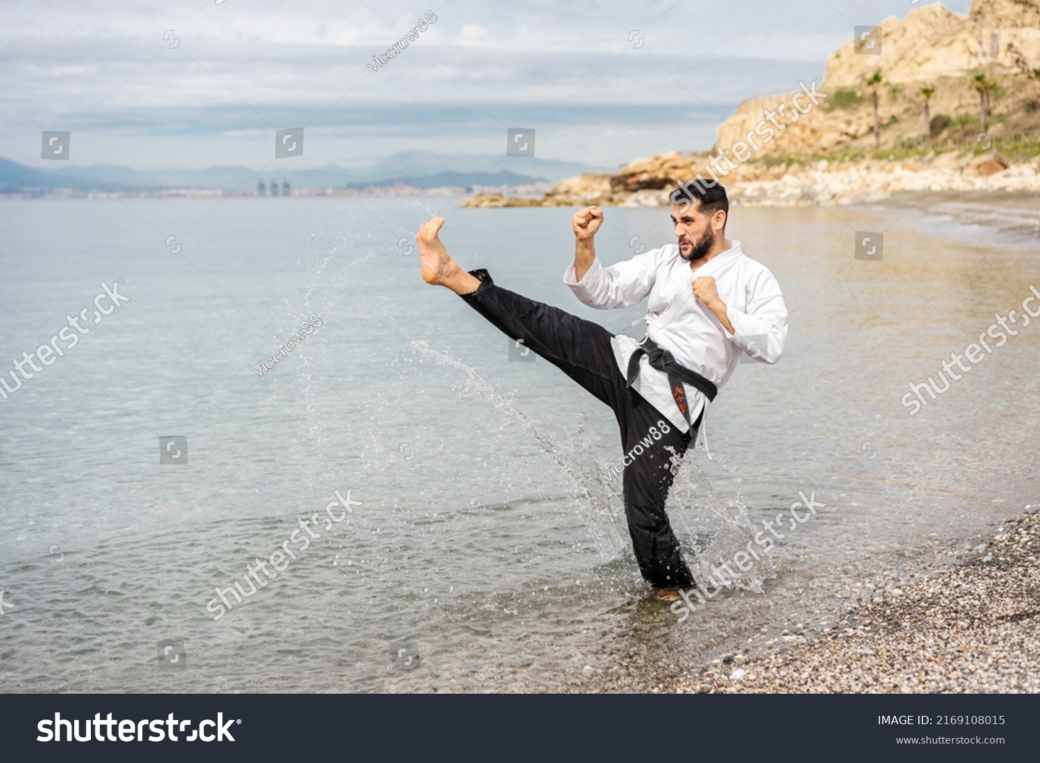 kung fu expert wearing a kimono and a black belt with the word "bushido" written in Japanese, performing a side kick on the shore of the beach splashing drops of water.  #2169108015