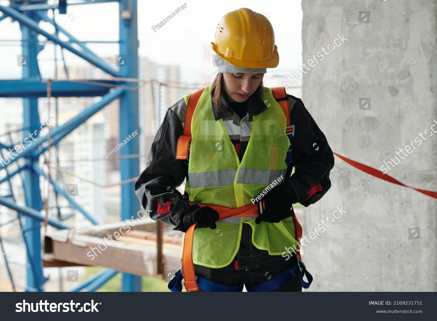 Young female builder in protective helmet and reflective vest fastening safety belt on waist before building work at construction site #2169231751
