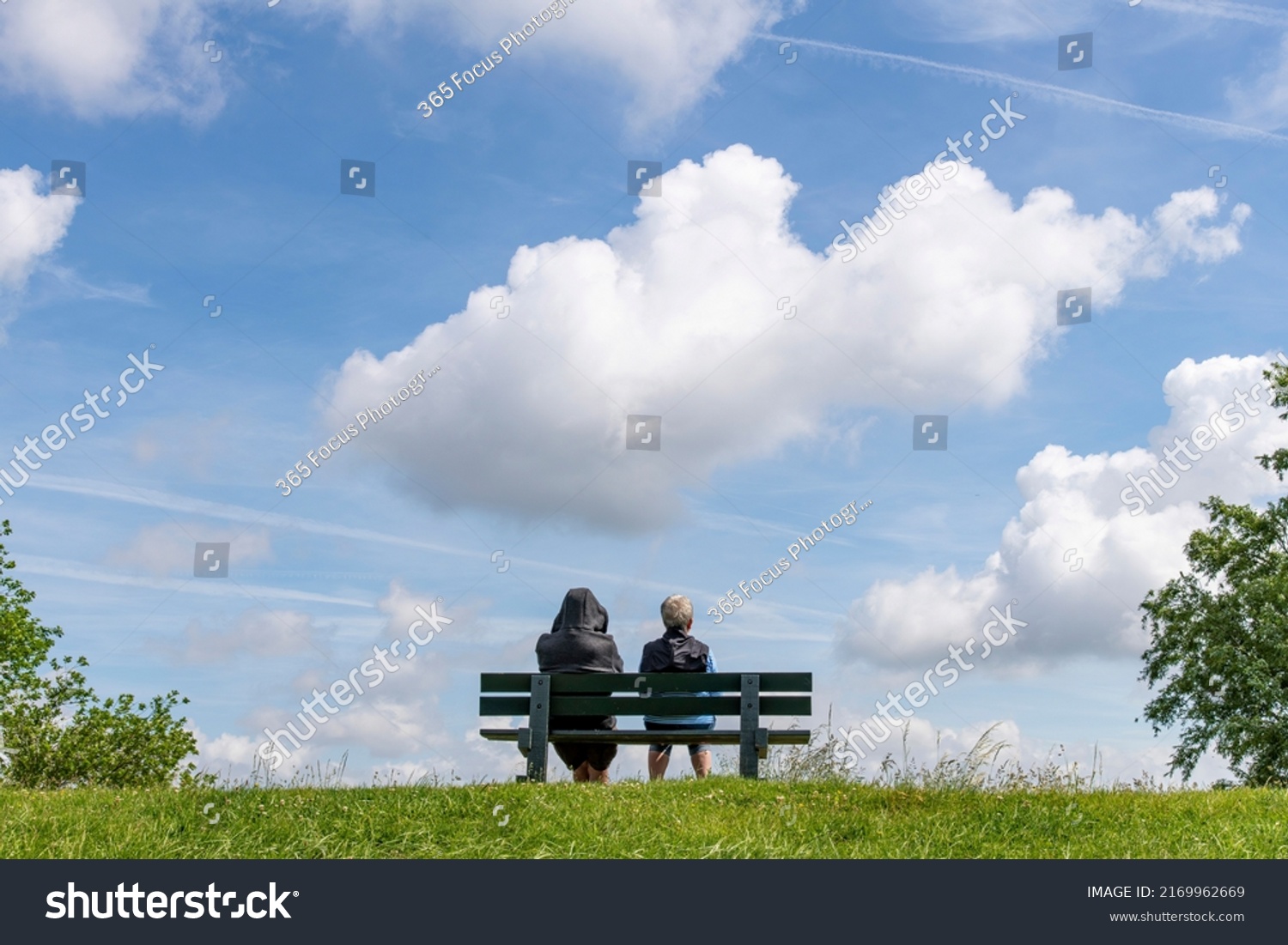 Low angle view of two people sitting on a park bench on top of a dike in the Netherlands looking in invisible distance with large cumulus clouds above in otherwise blue sky #2169962669