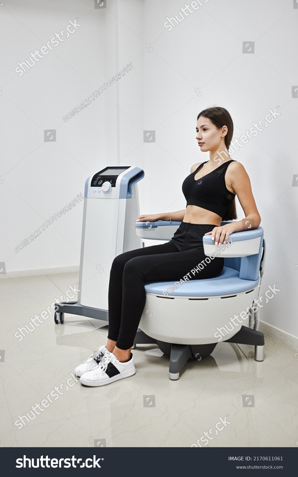 Young woman sitting on electromagnetic chair for stimulation of deep pelvic floor muscles and restoring neuromuscular control at the clinic #2170611061
