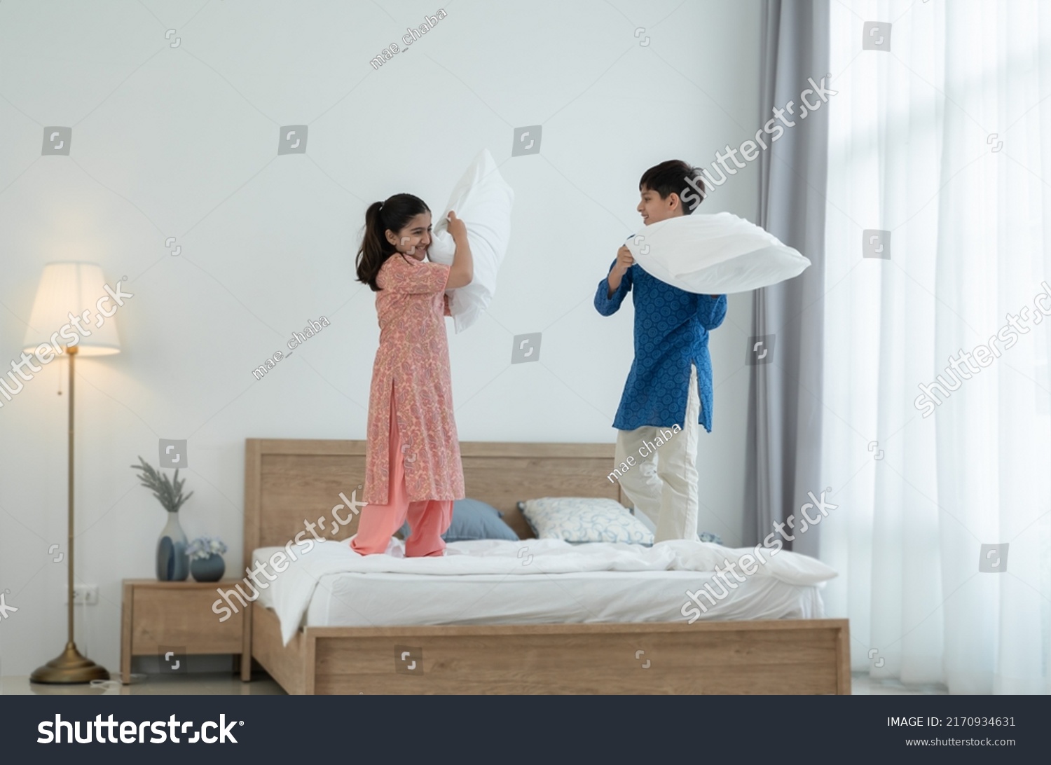 Two happy Indian brother and sister in traditional clothing standing on bed, playing pillow fight, having fun together at home. Playful kids, Siblings relationship concept #2170934631