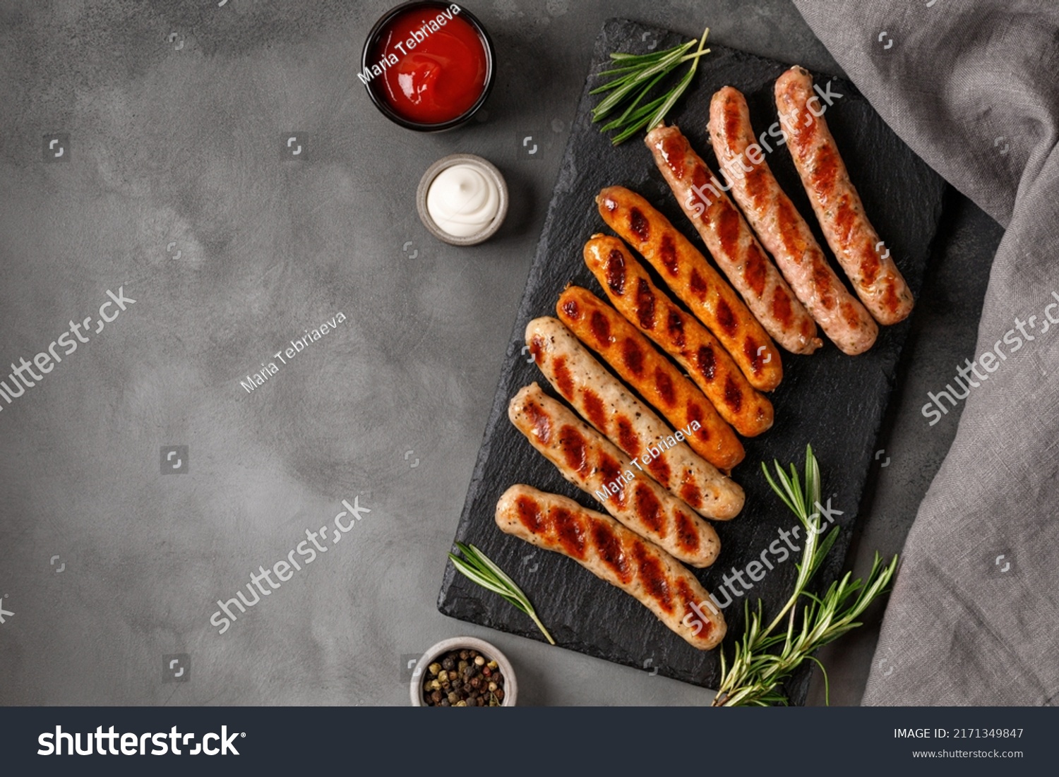 Assorted grilled sausages on barbecue with sauces. Assortment of different sausages on slate board and dark background. Top view, copy space. #2171349847