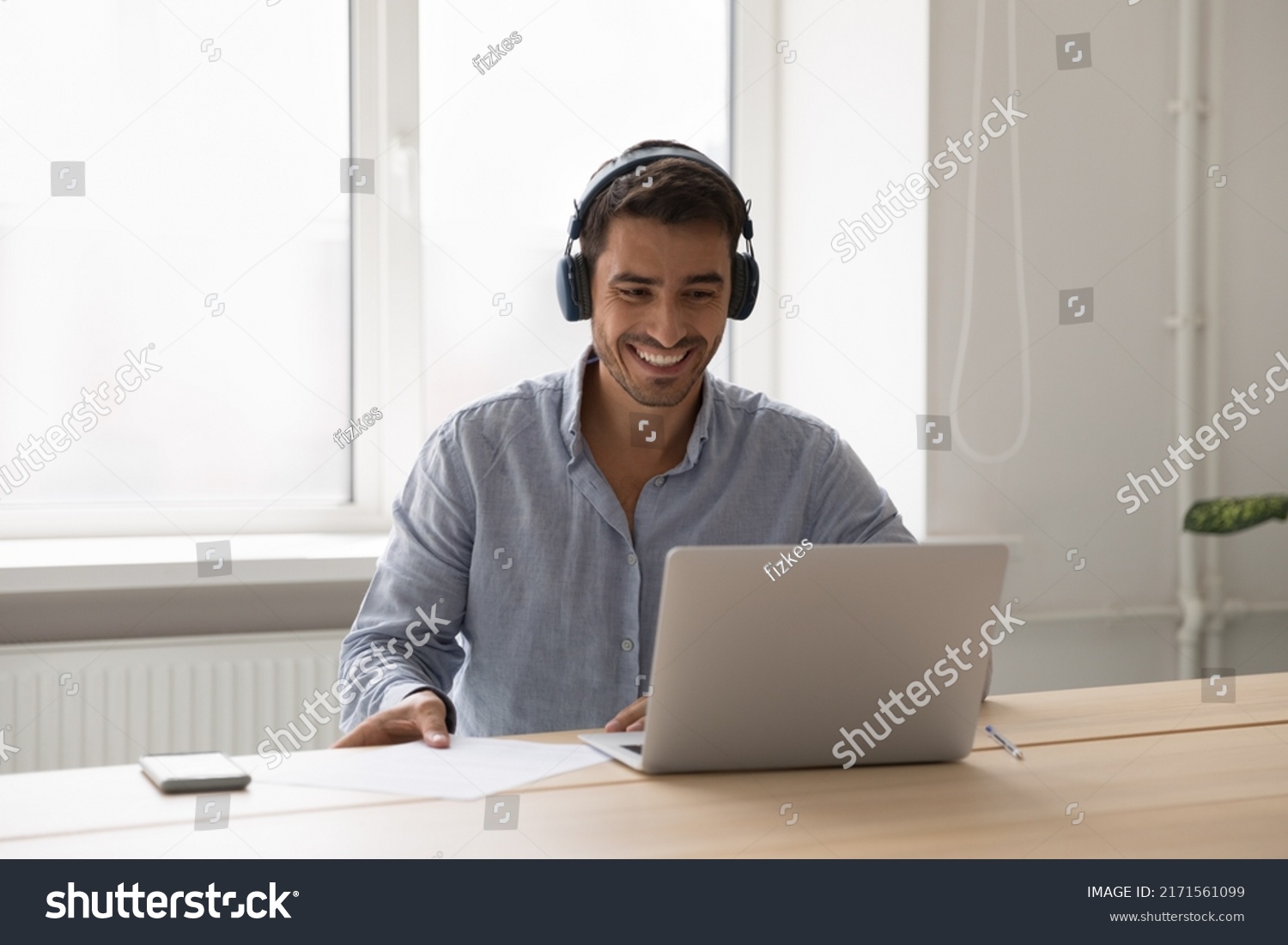 Man in headphones sit at desk with laptop working, take part in briefing on video call app. Millennial guy listens audio course or studying use modern tech. Virtual event, on-line counselling concept #2171561099