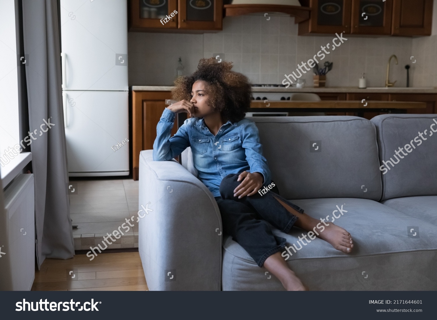 Sad African teen girl sit on sofa lost in thoughts, thinks staring into distance, looks upset experiences first unrequited love, having low self-esteem, problems, feels insecure. Teen problem concept #2171644601