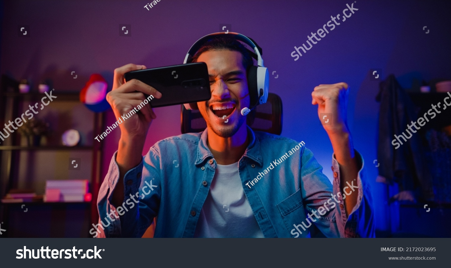 Happy asia man gamer wear headphone competition play video game online with smartphone colorful neon light in living room at night modern house. Esport streaming game online, Home quarantine activity. #2172023695