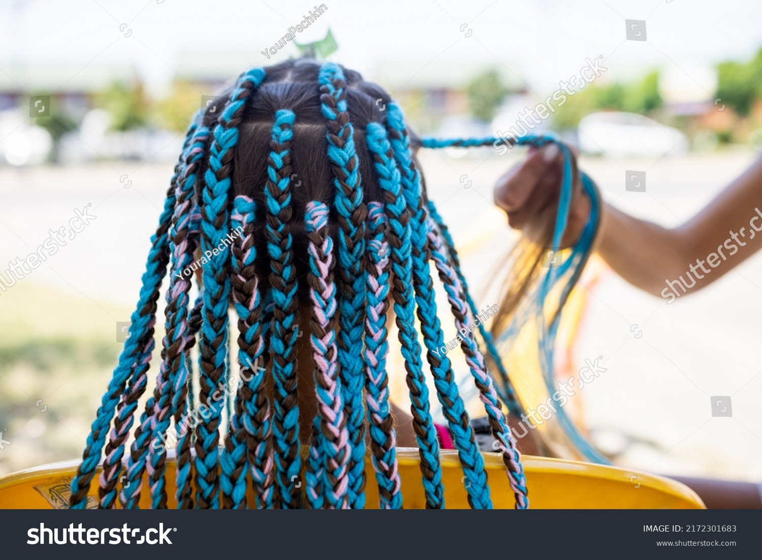 A little waiting tanned girl in a bright yellow summer suit, weaves colored pink-blue African braids in her dark wet thick hair on a hot sunny sea day #2172301683