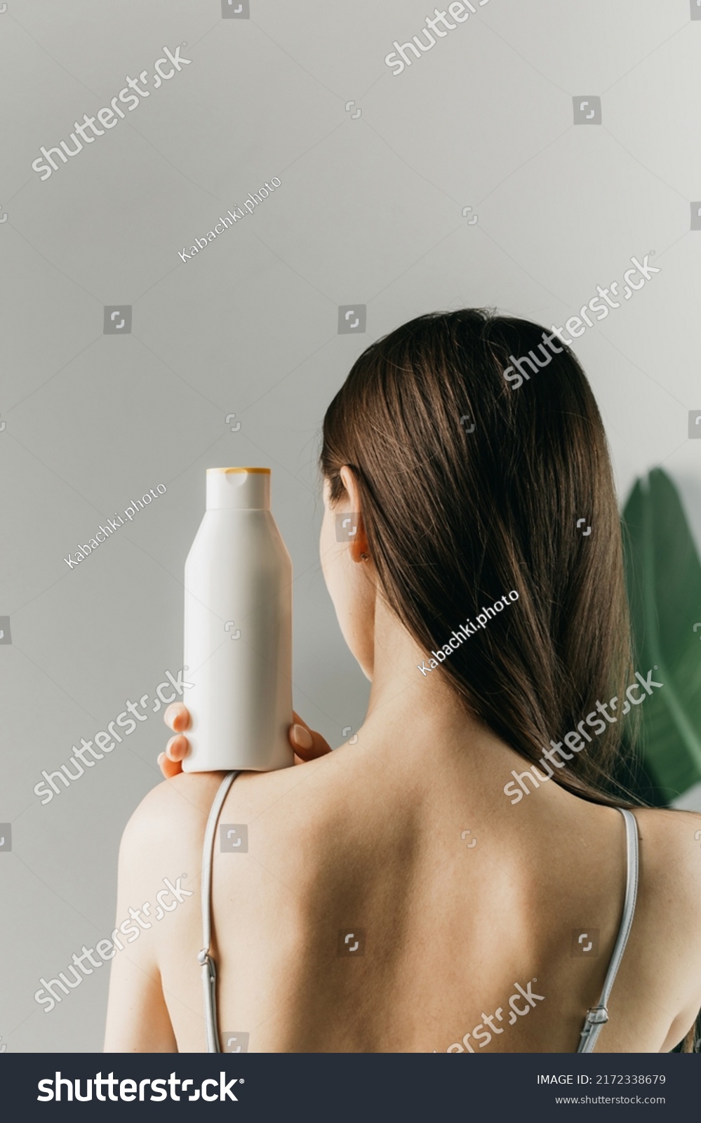 Woman holds a white bottle of cosmetics in her hand, standing with her back to the camera. Mock up shampoo, gel. #2172338679