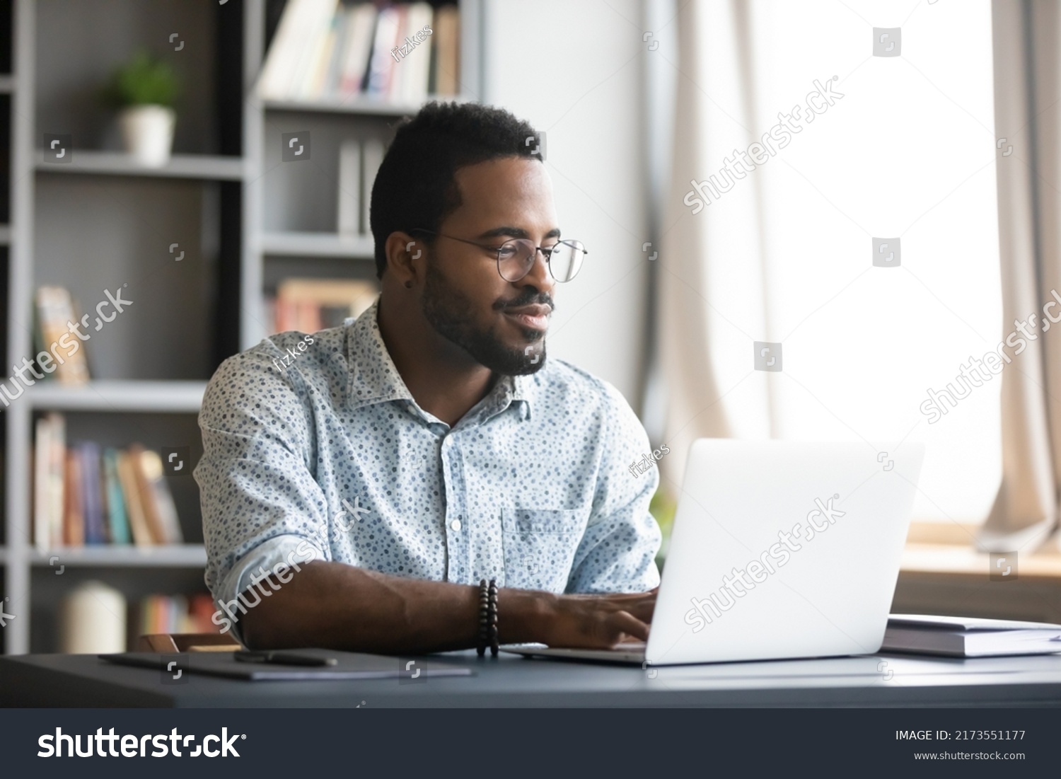 Satisfied African American man wearing glasses looking at computer screen, reading good news in email, chatting in social network with friends, freelancer blogger working on online project #2173551177