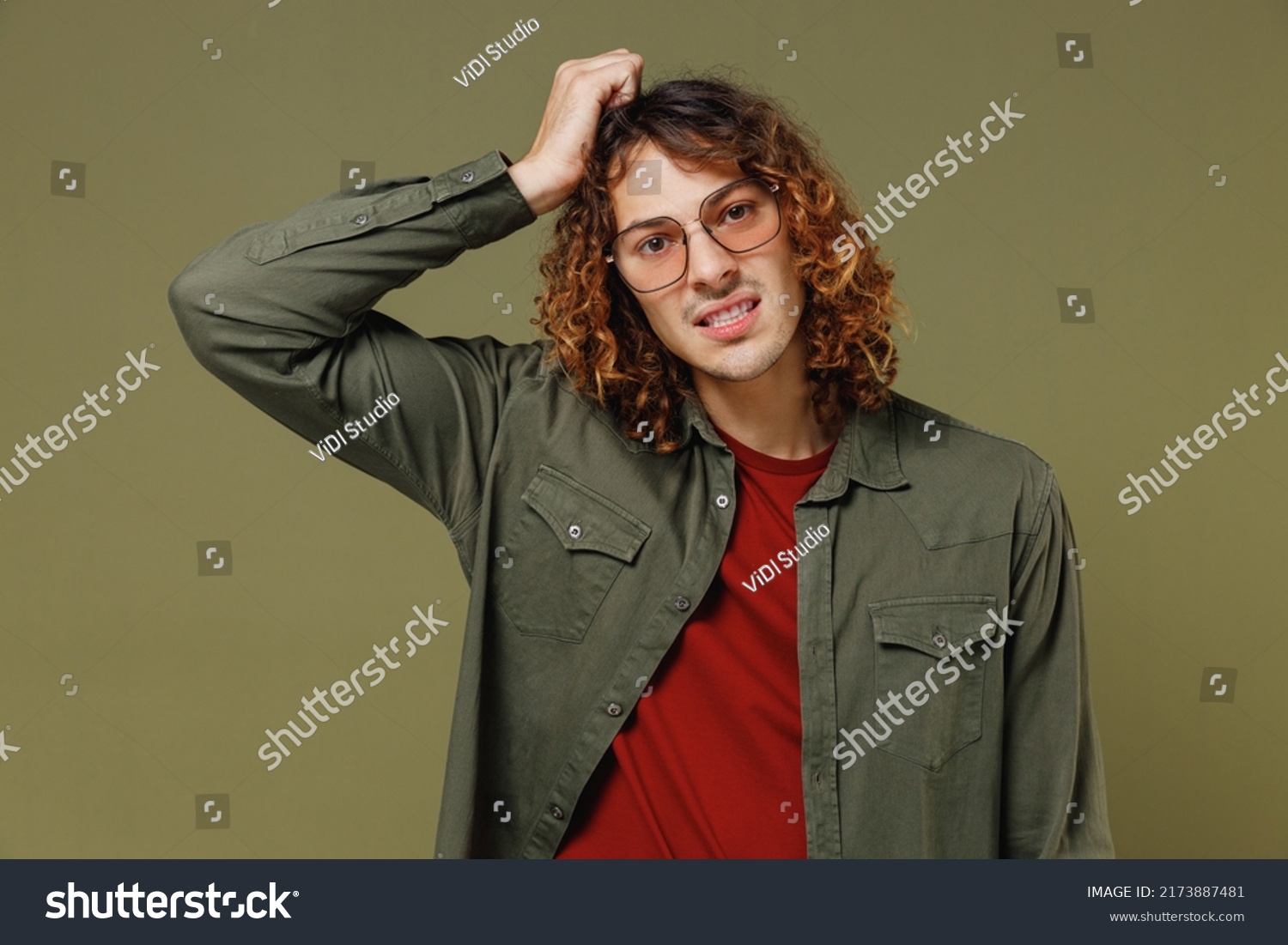 Confused disconcerted unnerved aggrieved upset young brunet curly man 20s wears khaki shirt glasses looking camera put hands on head scratching isolated on plain olive green background studio portrait #2173887481