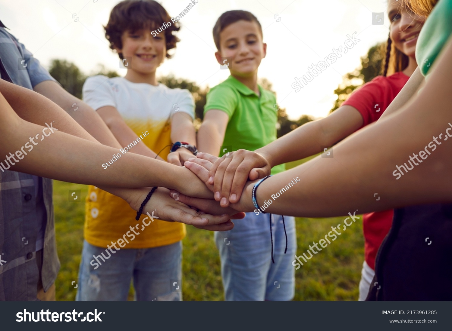 Bunch of children playing together on a good summer day. Group of happy friends standing in a circle in a green park or field, smiling and stacking their hands. Cropped closeup shot. Teamwork concept #2173961285