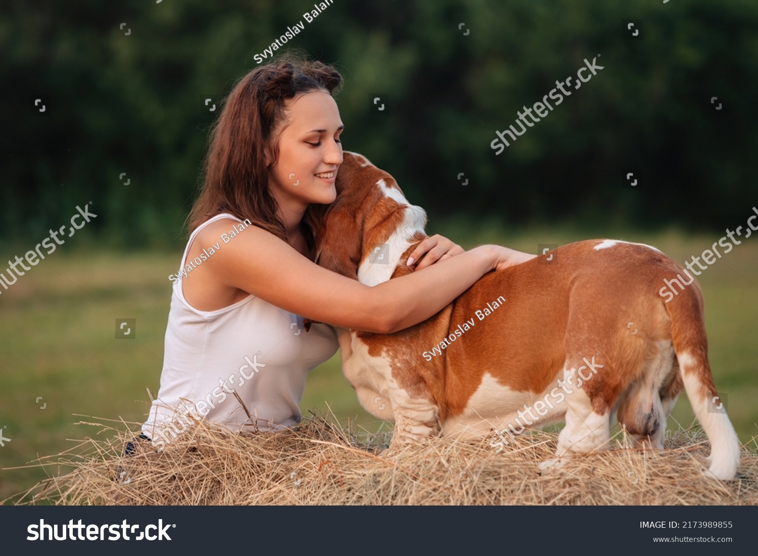 A young adult girl walks with a Basset Hound dog in nature. The owner hugs the pet. #2173989855