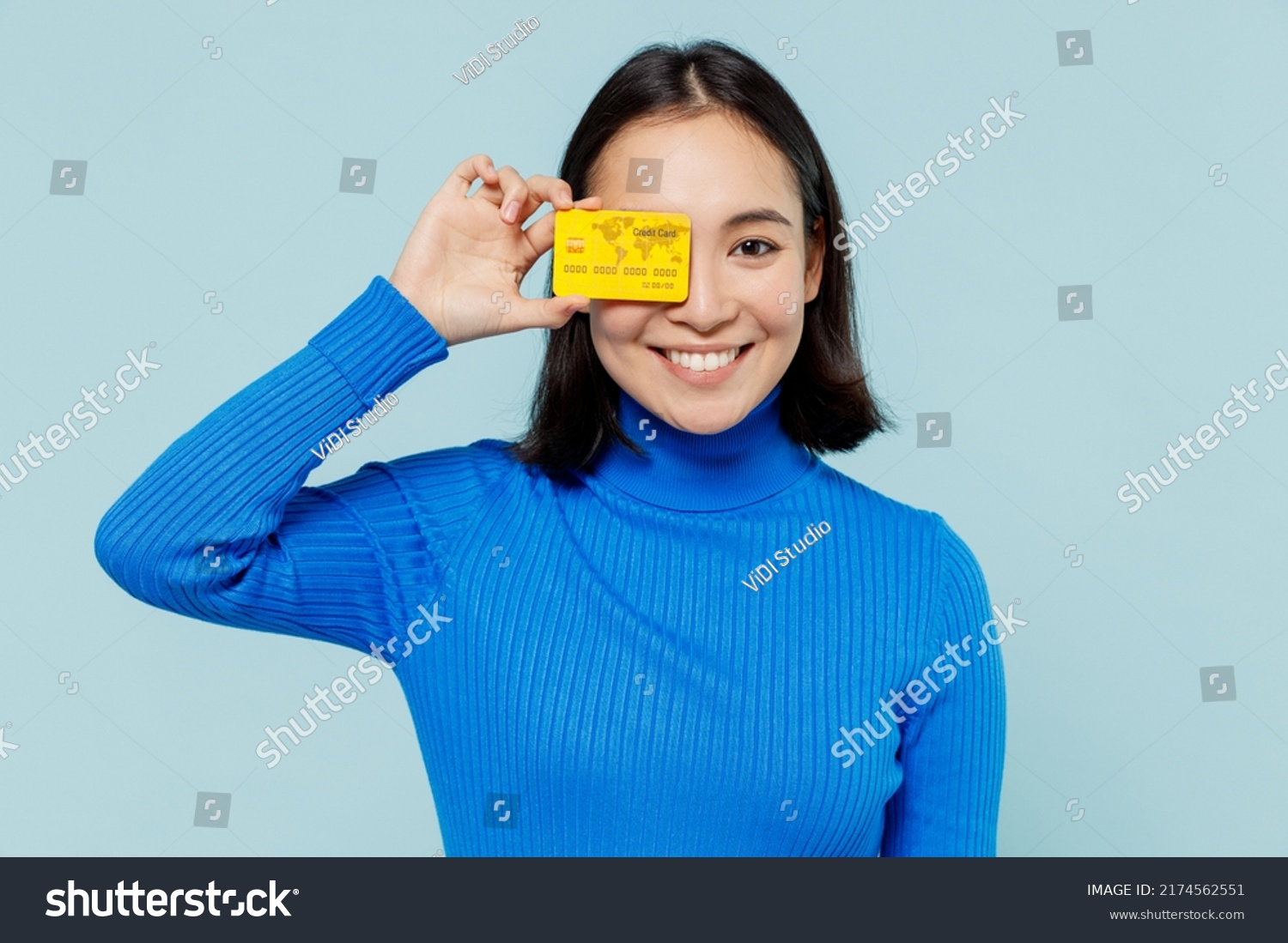 Fascinating fun joyful young woman of Asian ethnicity 20s years old wears blue shirt hold in hand credit bank card cover close hiding eye isolated on plain pastel light blue background studio portrait #2174562551