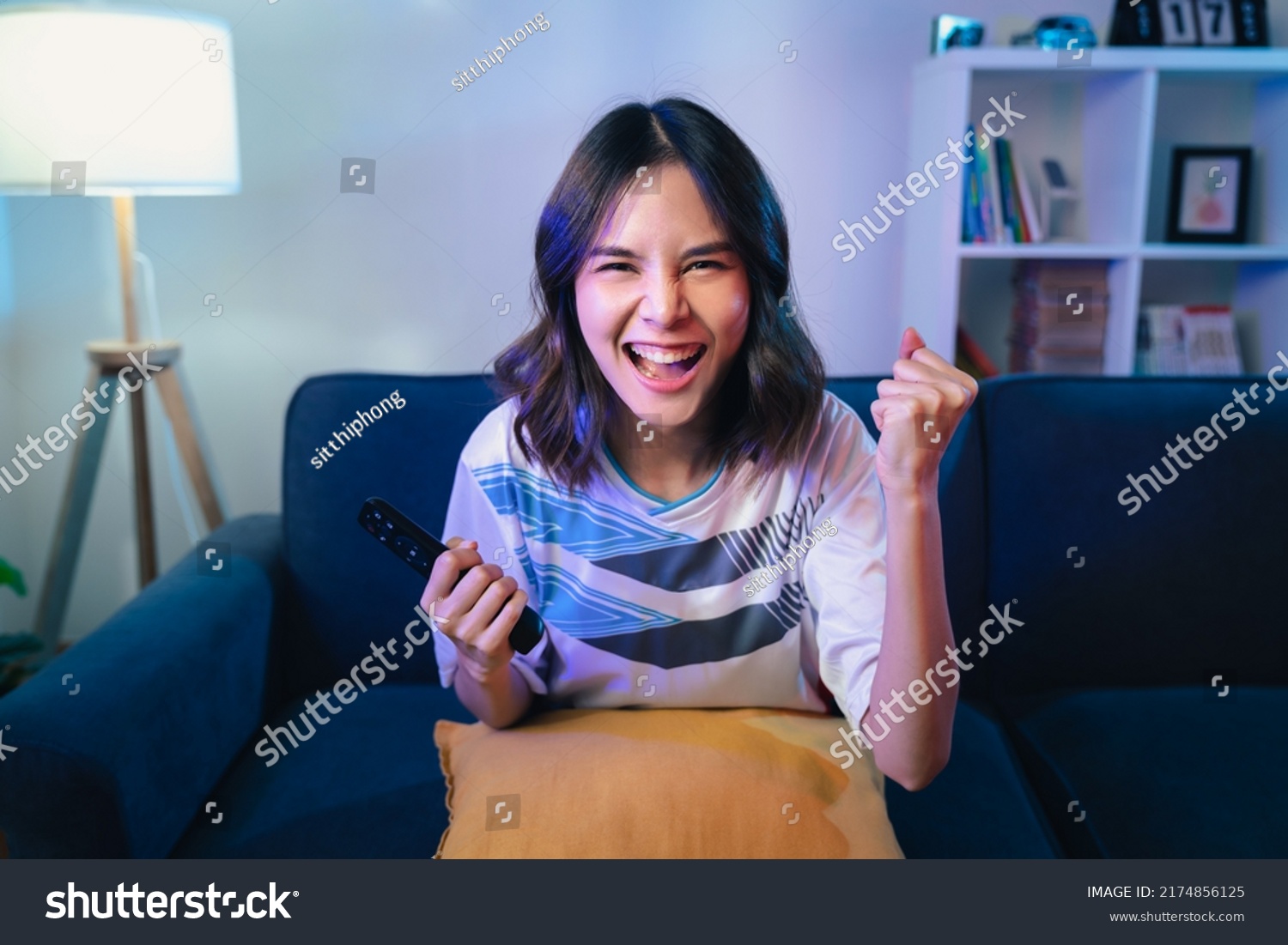 Excited young Asian woman watching TV and cheering sport games competition with laugh victory on sofa at home. #2174856125