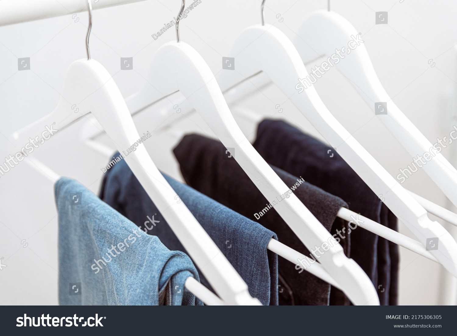 Many blue denim jeans hanging on white clothes hangers on clothing rack. Close up of folded casual denim jeans in wardrobe with coat hangers #2175306305