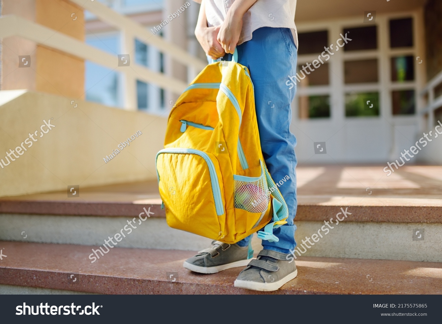 Little student with a backpack on the steps of the stairs of school building. Close-up of child legs, hands and schoolbag of boy standing on staircase of schoolhouse. Kids back to school concept. #2175575865