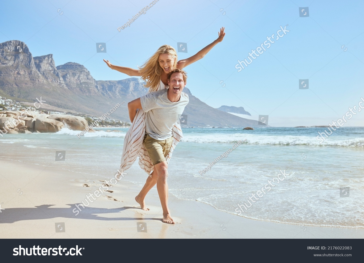 Portrait of a happy and loving young couple enjoying a day at beach in summer. Cheerful affectionate husband giving his joyful wife a piggyback while walking and having fun by the seashore on holiday #2176022083