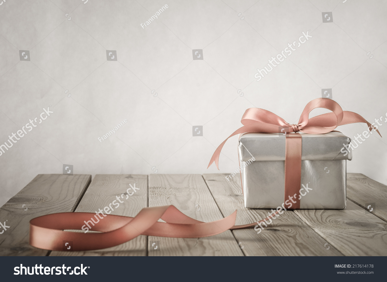 A gift box with closed lid, wrapped in silver paper and tied to a bow with a satin ribbon.  Placed on a weathered old wooden table with copy space behind and above. Cut ribbon remnant to the side.  #217614178