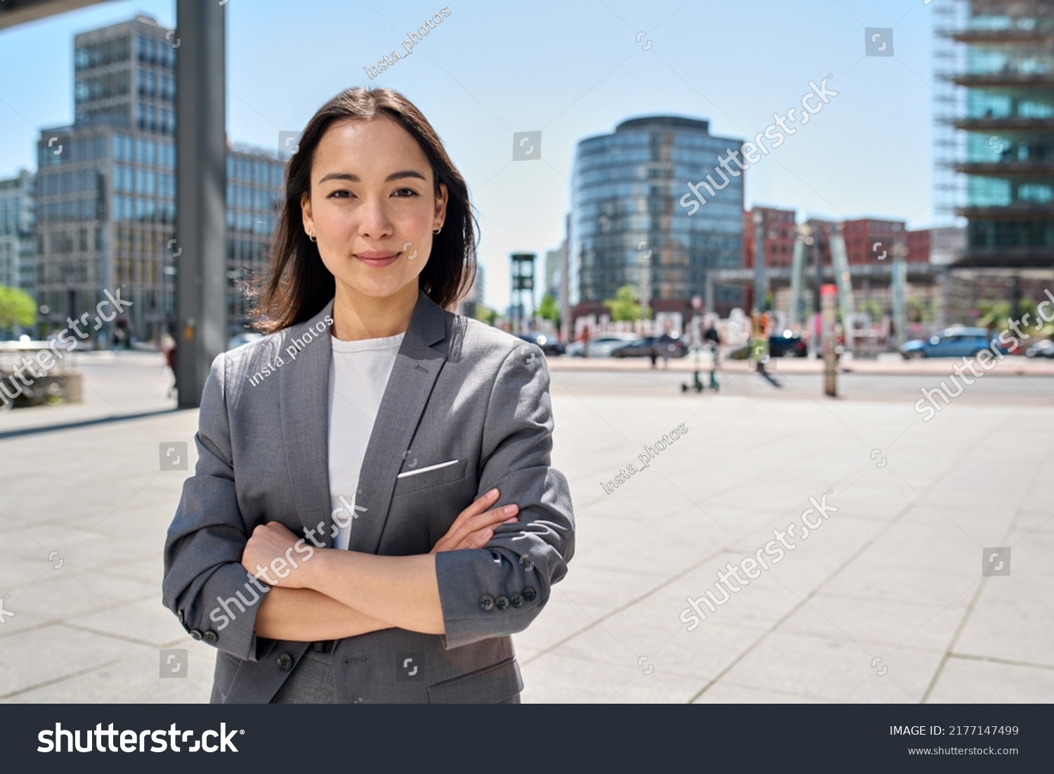 Young confident smiling Asian business woman standing on busy street, portrait. Proud successful female entrepreneur wearing suit posing with arms crossed looking at camera in big city outdoors. #2177147499