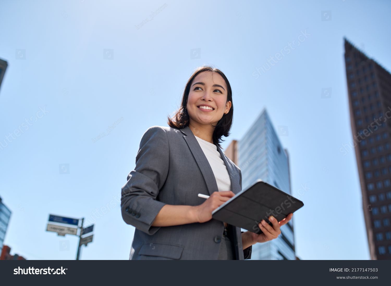 Smiling young Asian business woman leader entrepreneur, professional manager holding digital tablet computer using software applications standing on the street in big city on sky background. #2177147503