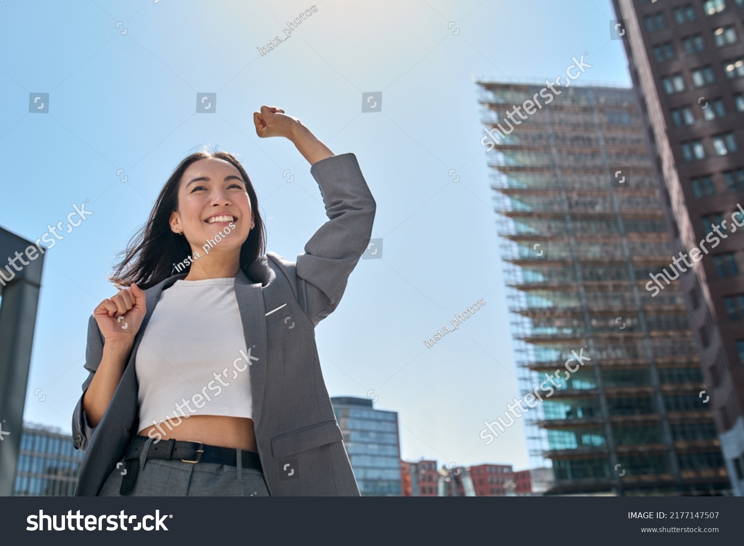 Young excited confident proud Asian business woman winner wearing suit standing on street, raising hands, feeling power, motivation, energy, celebrating career financial success in big city outdoors. #2177147507