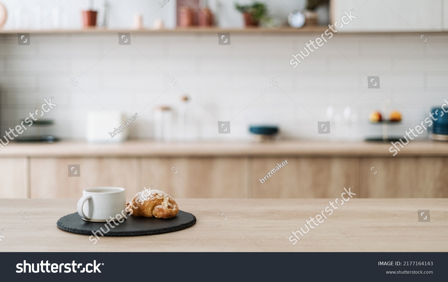 Croissant and coffee on kitchen countertop, against blurred minimalist interior with modern furniture. Selective focus at homemade pastry and tea drink in cup on wooden table, copy space, web banner #2177164143