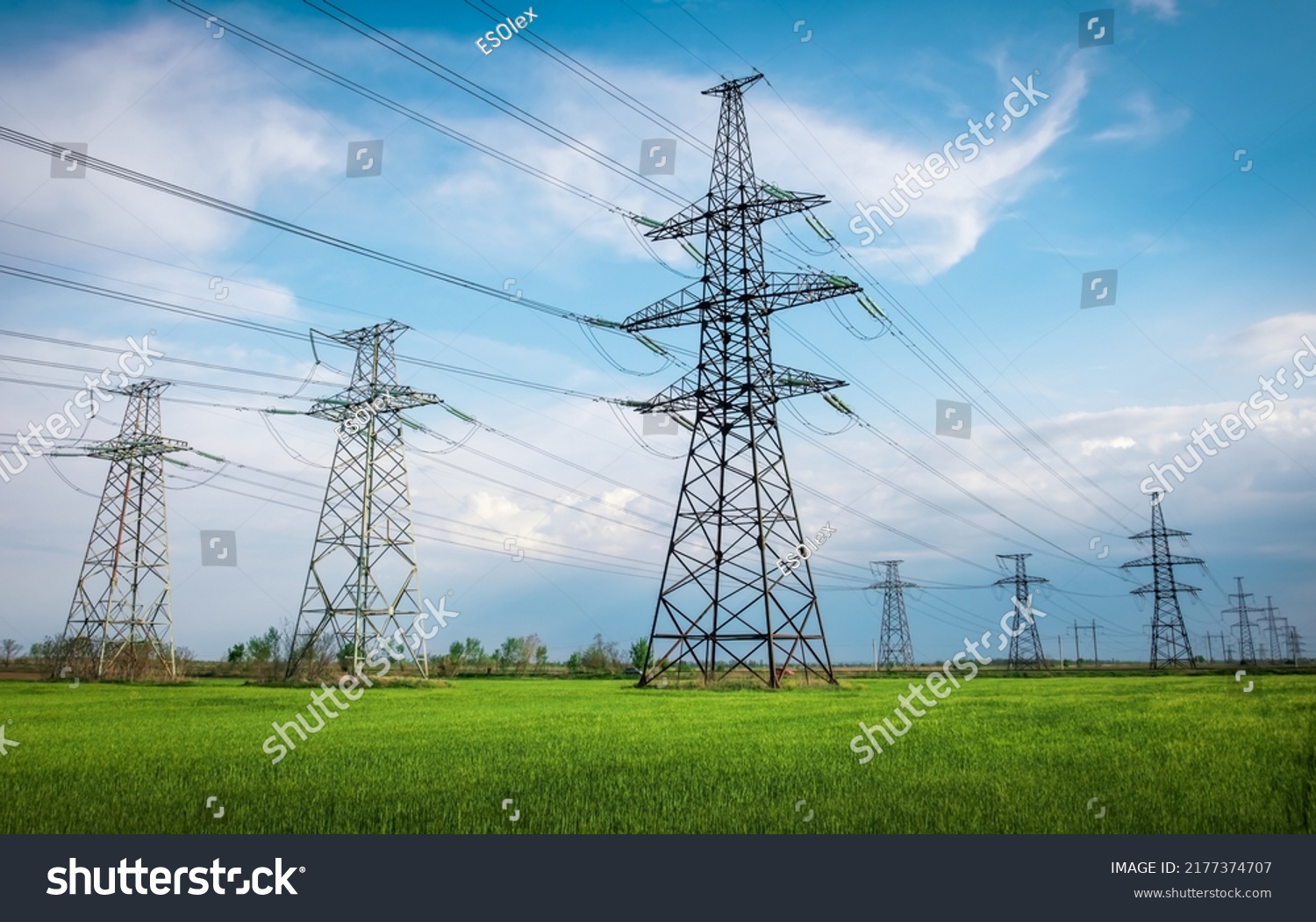 High voltage lines and power pylons in a flat and green agricultural landscape on a sunny day with clouds in the blue sky. Cloudy and rainy. Wheat is growing #2177374707