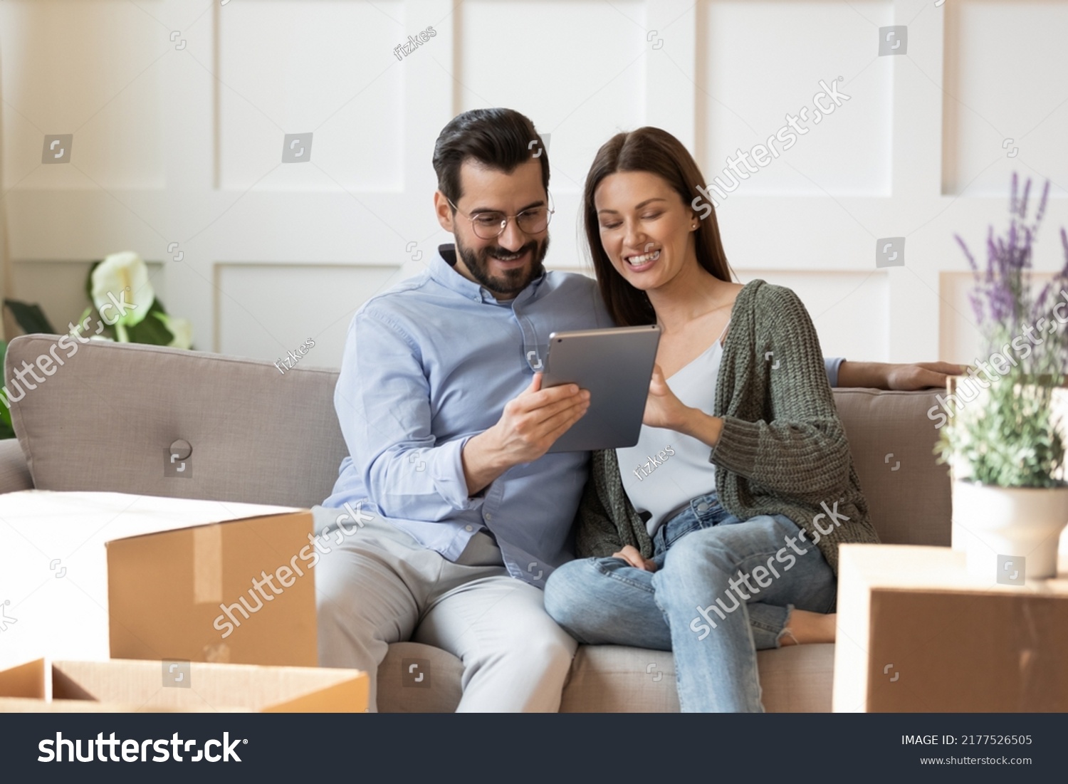 Smiling young Caucasian couple relax on sofa in living room on moving day watch video on tablet. Happy man and woman renters tenants rest on couch relocate to new home, use pad browsing internet. #2177526505