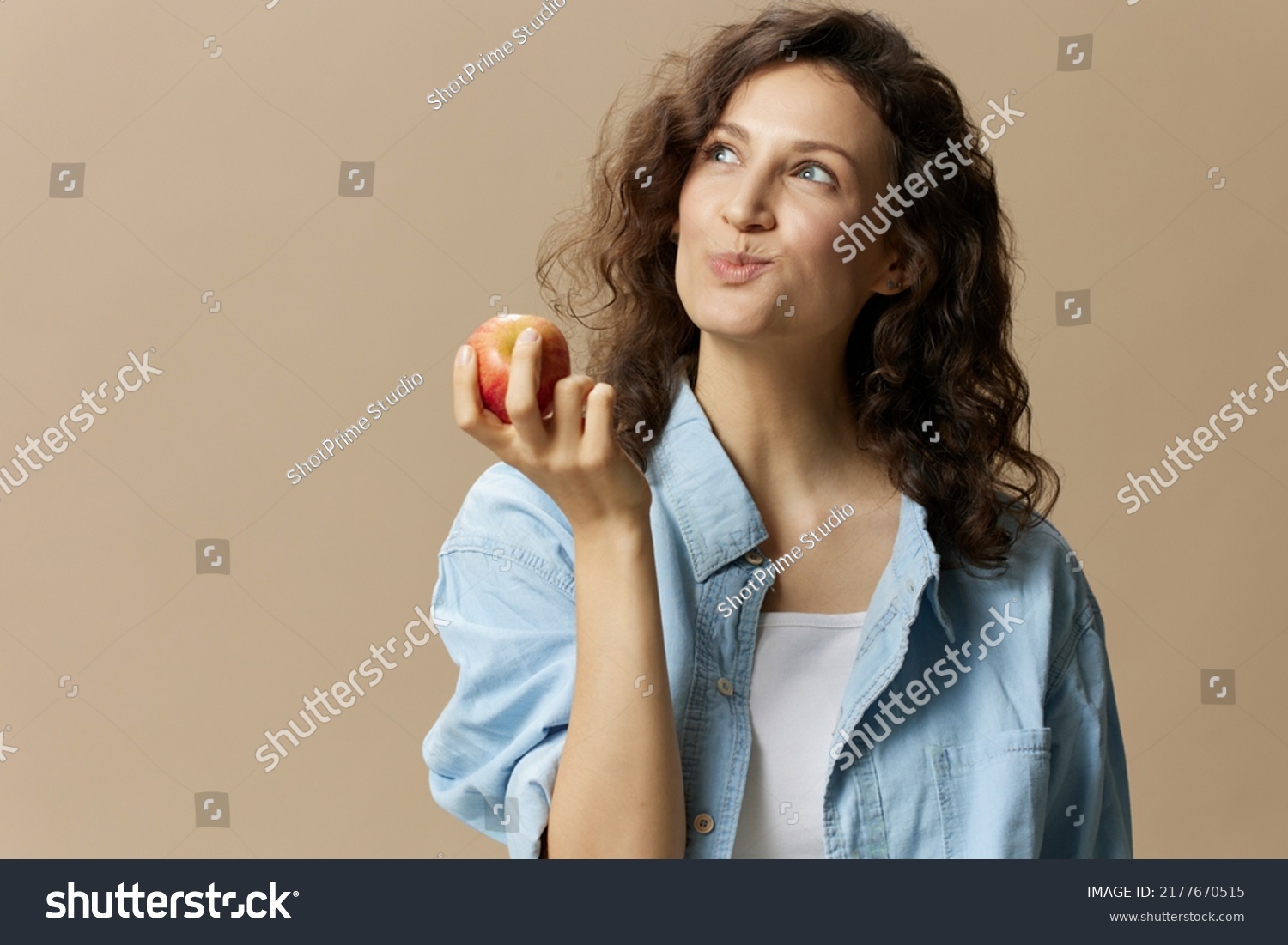 Funny happy cute curly beautiful female in jeans casual shirt chewing enjoy apple posing isolated on over beige pastel background. Healthy food. Natural eco-friendly products concept. Copy space #2177670515