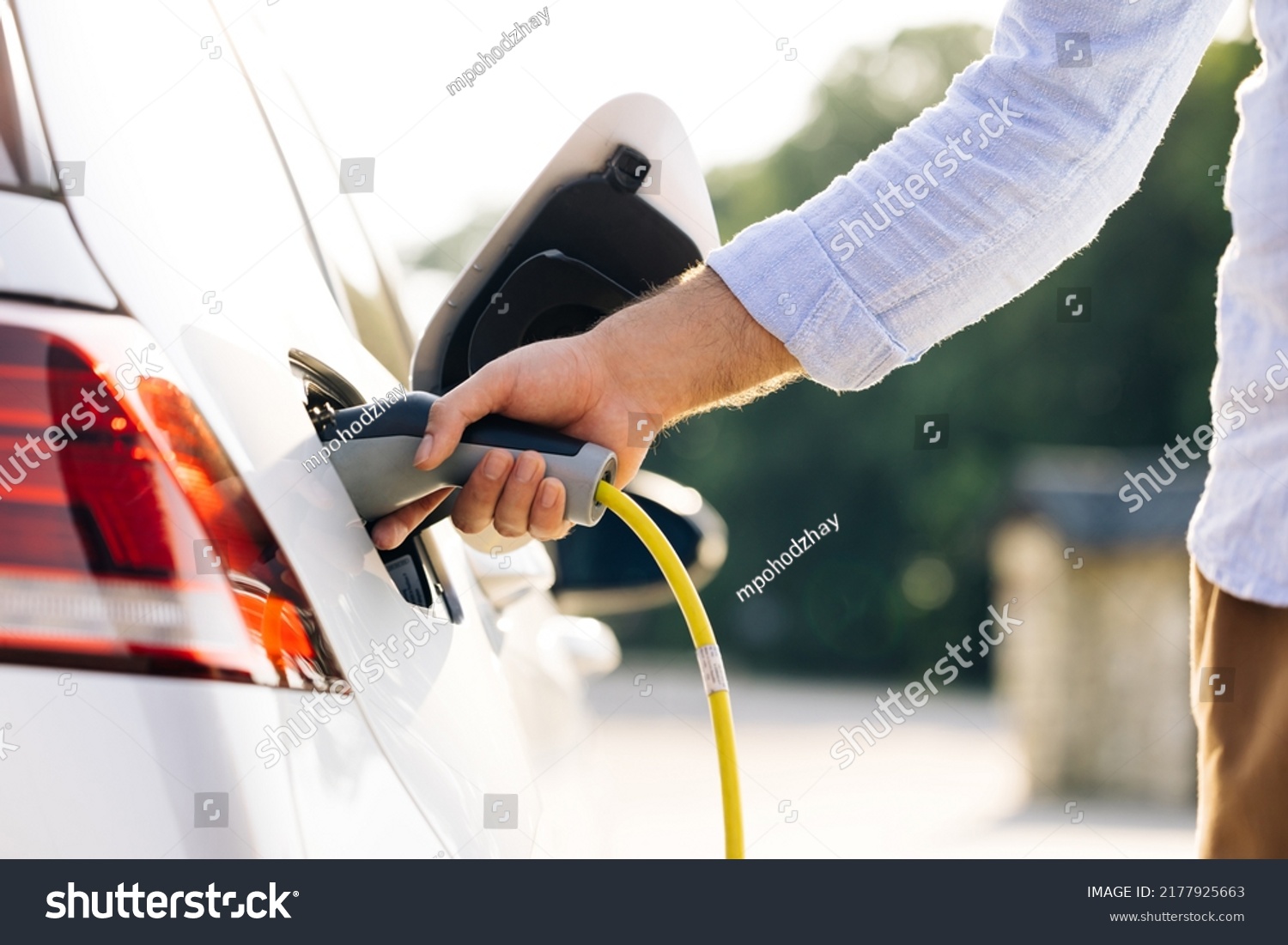 The driver of the electric car inserts the electrical connector to charge the batteries. Unrecognizable man attaching power cable to electric car. Electric vehicle Recharging battery charging port. #2177925663