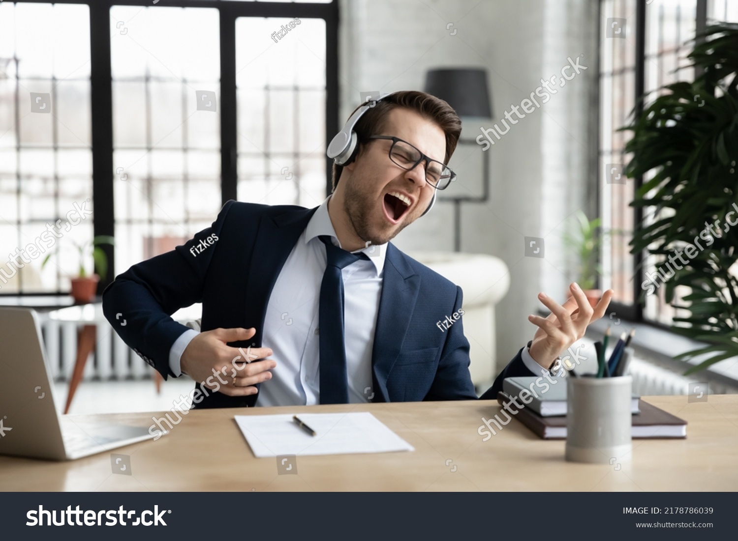 Funny overjoyed businessman wearing headphones pretending playing guitar, sitting at desk in modern office, excited employee executive singing, listening to favorite music, having fun during break #2178786039